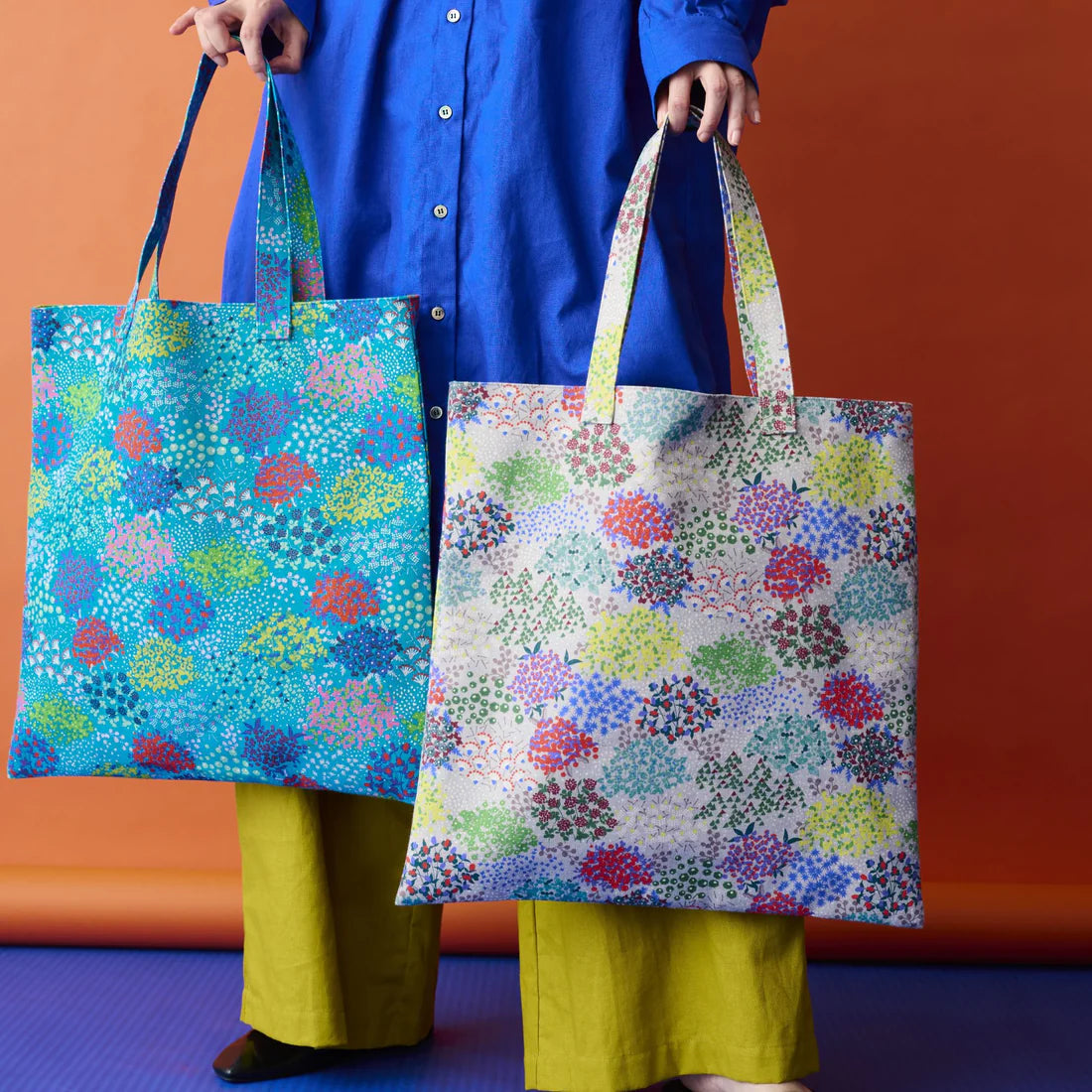 A person is holding two colorful tote bags with floral patterns against an orange background.